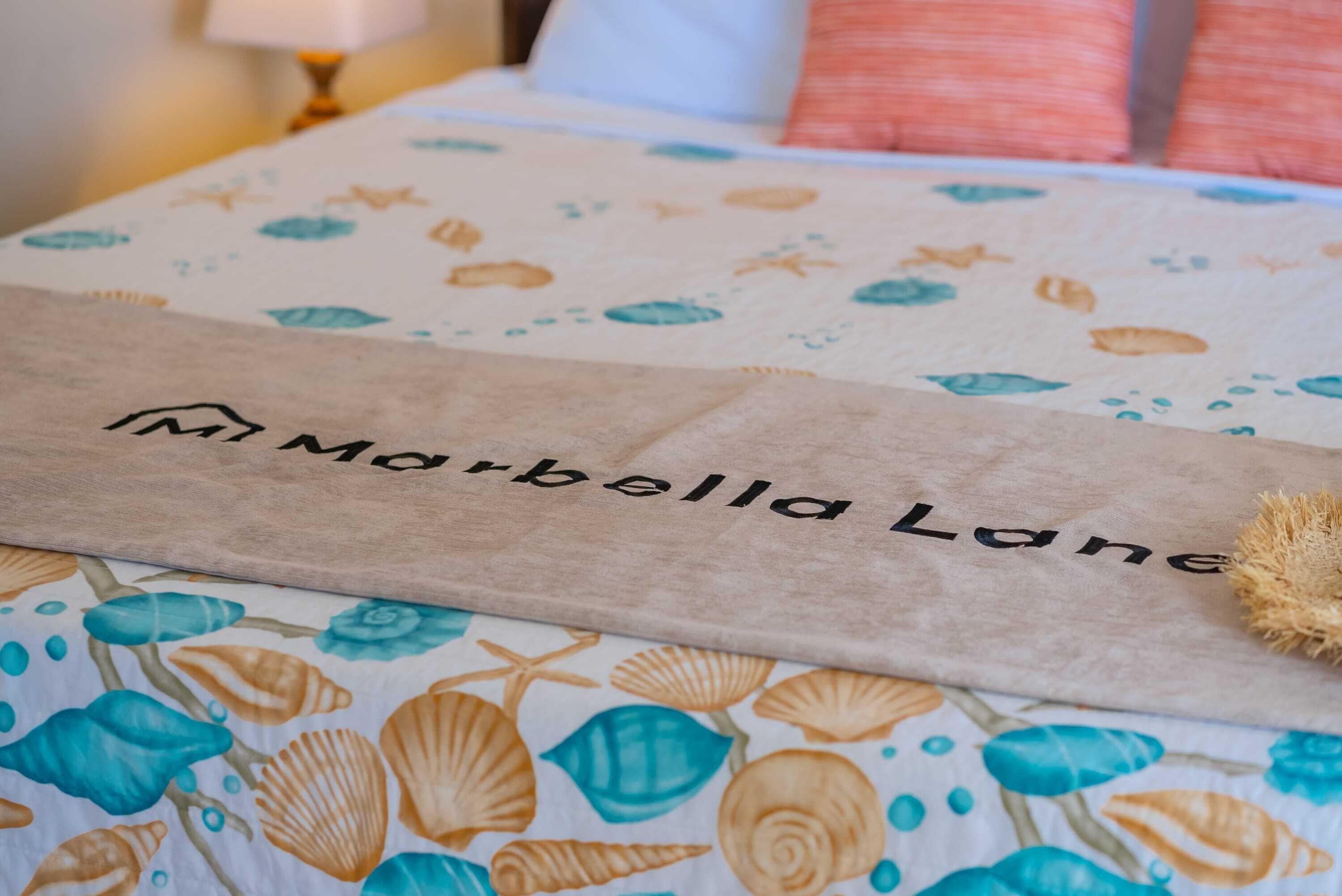 A bed with a floral-patterned bedspread and a beige towel with 'Marbella Lane' text.