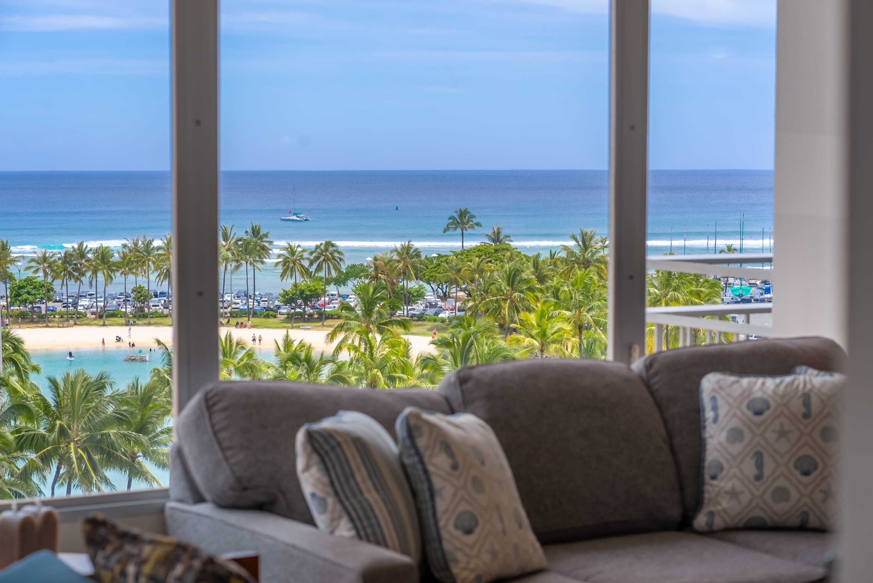 A room with a large window overlooking a beach with palm trees and a pool.