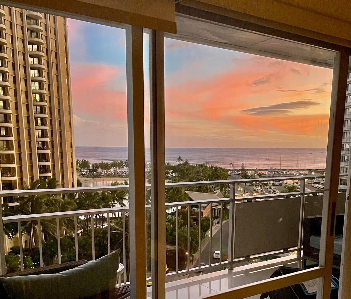 Un balcon offrant une vue sur un coucher de soleil sur l’océan, des palmiers et un immeuble de grande hauteur.