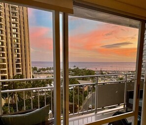 A balcony with a view of a sunset over the ocean, palm trees, and a high-rise building. - @ Marbella Lane - Cozy 2BR w/ Amazing Ocean Views (Honolulu)