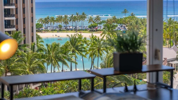 Vista desde un balcón sobre una playa con palmeras, una piscina y el océano.