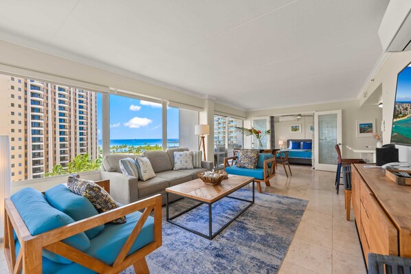 A spacious living area with a large window overlooking the ocean, a sofa set, a coffee table, and a dining area with chairs. - @ Marbella Lane - Cozy 2BR w/ Amazing Ocean Views (Honolulu)