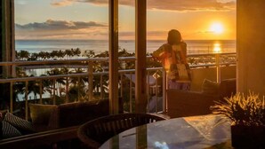 Un balcón con vistas a la puesta de sol sobre el océano, palmeras y un paisaje urbano lejano.