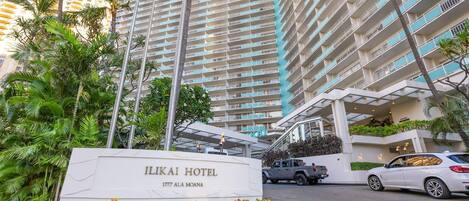 L’entrée de l’hôtel Ilikai, avec une enseigne, est entourée de palmiers et donne sur un parking où sont garées des voitures.