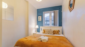 Ein Schlafzimmer mit Bett, Fenster, Nachttisch, Lampe und einem an der Wand befestigten Spiegel.
