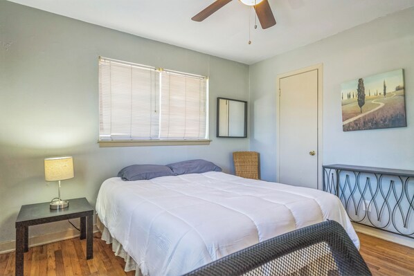 Un dormitorio con cama, una mesita de noche con lámpara, una ventana con persianas, una puerta y un cuadro en la pared.
