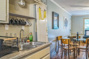 Una cocina con encimeras de granito, un lavamanos de acero inoxidable y un tendedero para ollas y sartenes. Hay una zona de comedor con una mesa de vidrio y sillas de madera.