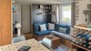 A cozy living room with a sofa, a coffee table, and a dining table. There are shelves with decorative items and a window with curtains.