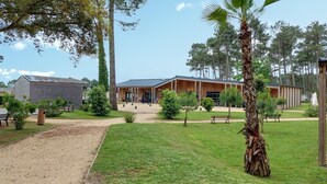 A building with a covered patio area, a dirt path leading to the entrance, and a grassy lawn with trees and shrubs.