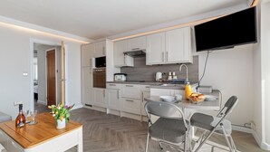 A modern kitchen with white cabinets, a flat-screen TV mounted above the counter, and a dining area with a wooden table and metal chairs. - 9 Strandways - Sea Views, Parking, Beach Access (Saundersfoot)