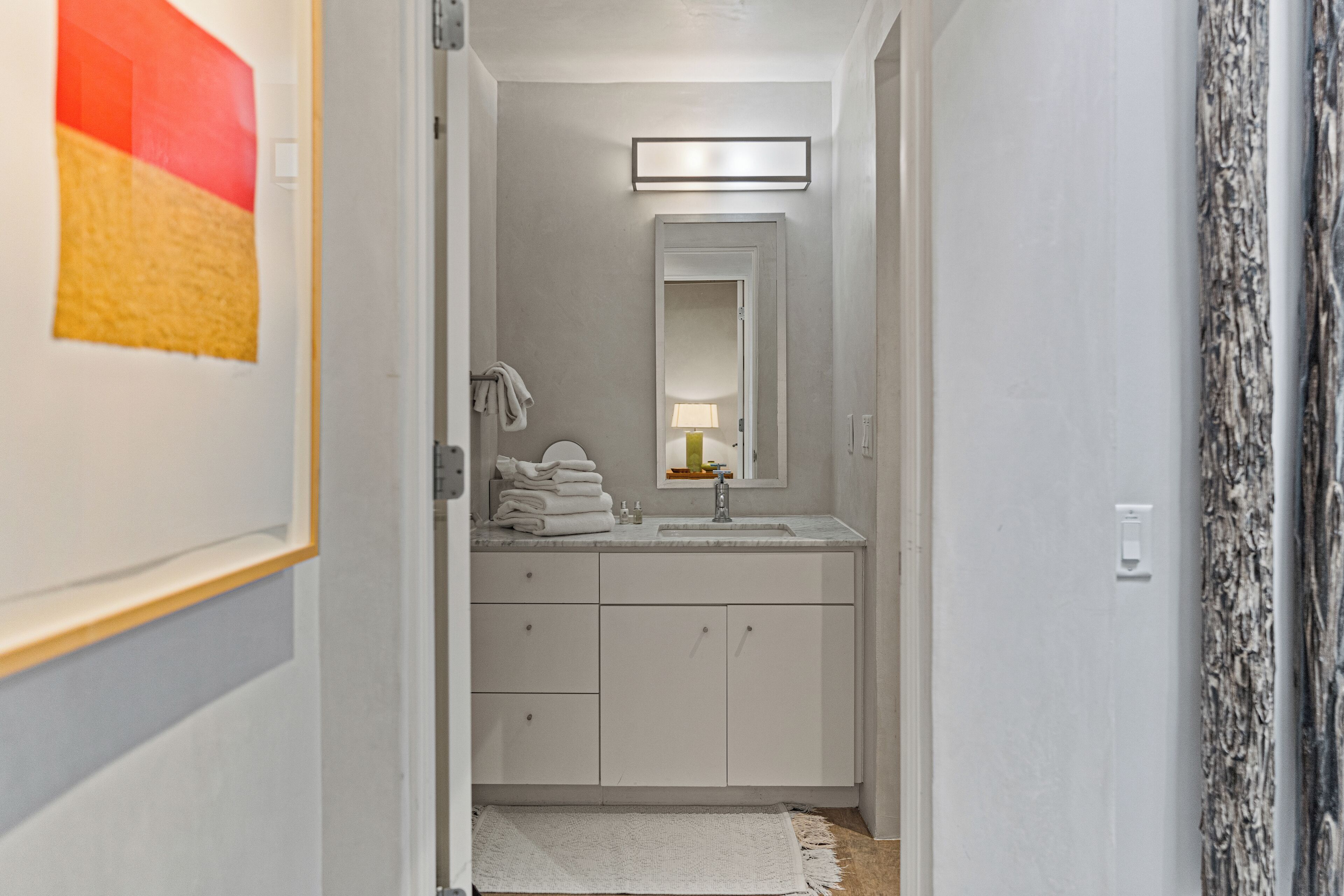 Un couloir étroit mène à une salle de bain équipée d’un lavabo blanc, d’un miroir et d’un porte-serviettes.