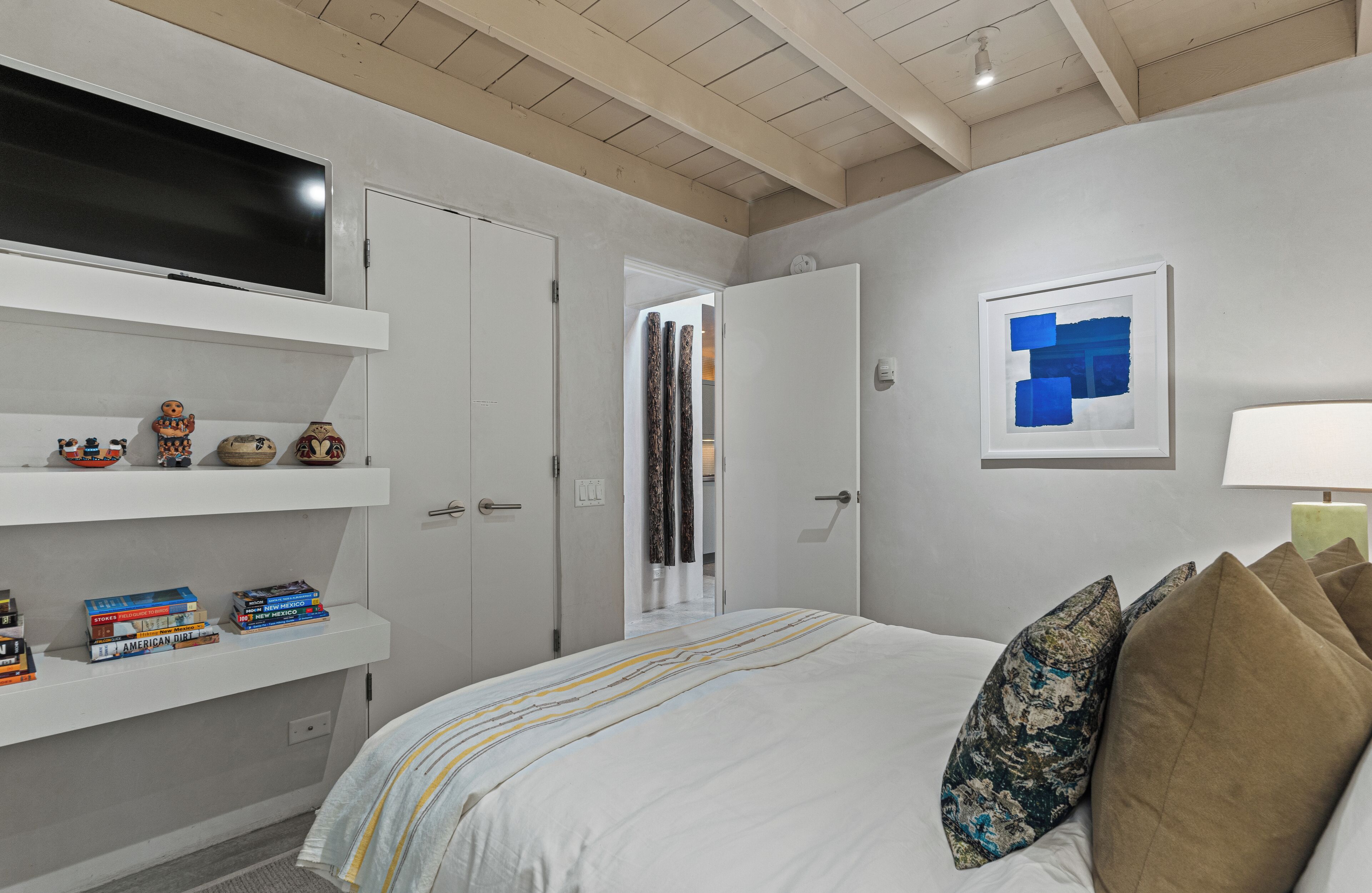 A bedroom with a bed, a TV mounted on the wall, a shelf with books, and a door leading to another room.