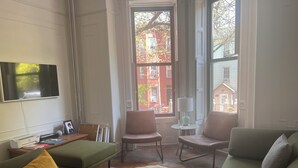 Ein Wohnzimmer mit einem grünen Sofa, zwei Sesseln, einem kleinen Tisch mit einer Lampe, einem an der Wand montierten Fernseher und Blick auf ein rotes Backsteinhaus durch ein Fenster.