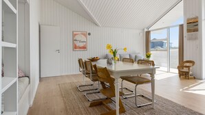 A dining area with a white table, wicker chairs, and a view of the beach through large windows. - Lovely home in Stege with sauna (Stege)
