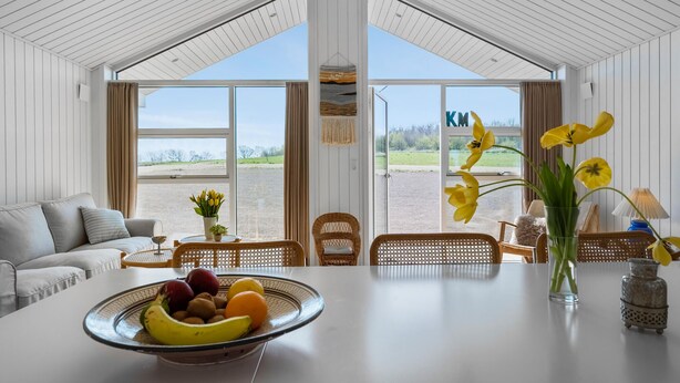 A spacious room with a high ceiling, large windows, a dining table with a fruit bowl, wicker chairs, and a view of a field. - Lovely home in Stege with sauna (Stege)