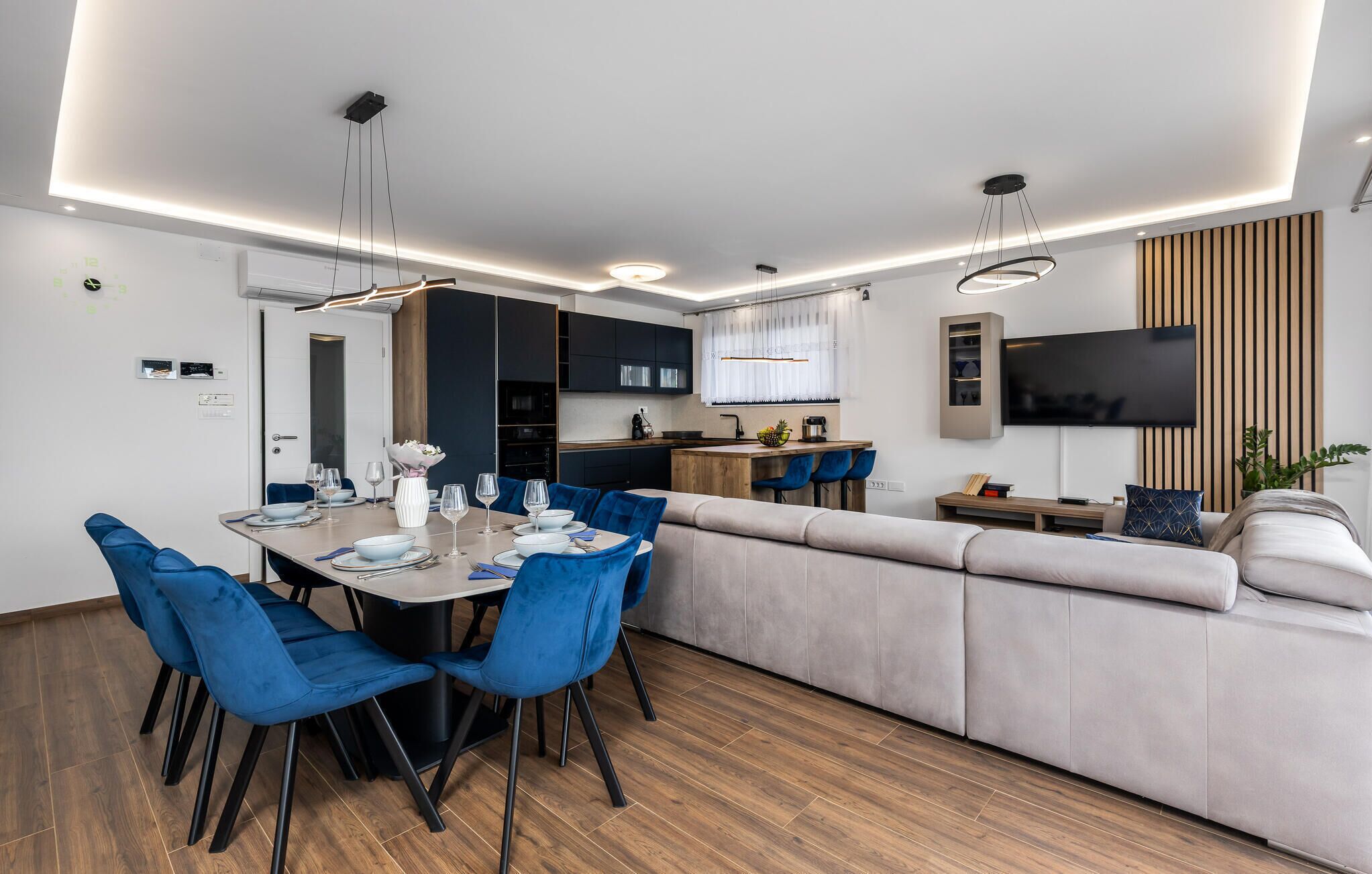A modern living room with a sofa, dining table, and chairs. There is a kitchen area with a TV and a plant.