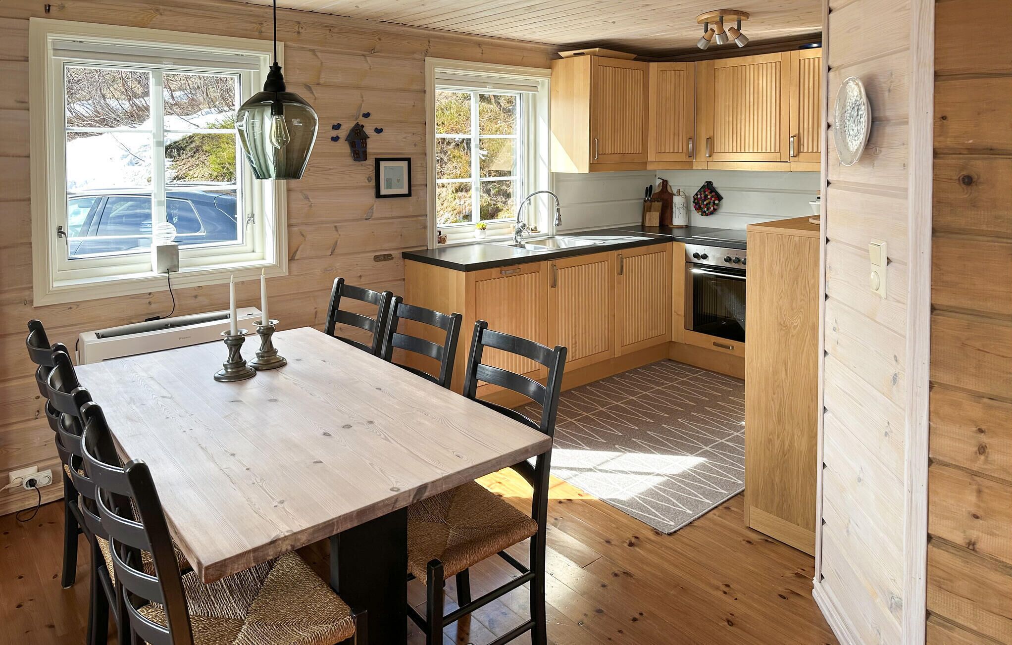 A wooden interior with a dining table, chairs, a kitchen area with wooden cabinets, a stove, and a window with a view of the outside.