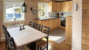 A wooden interior with a dining table, chairs, a kitchen area with wooden cabinets, a stove, and a window with a view of the outside.