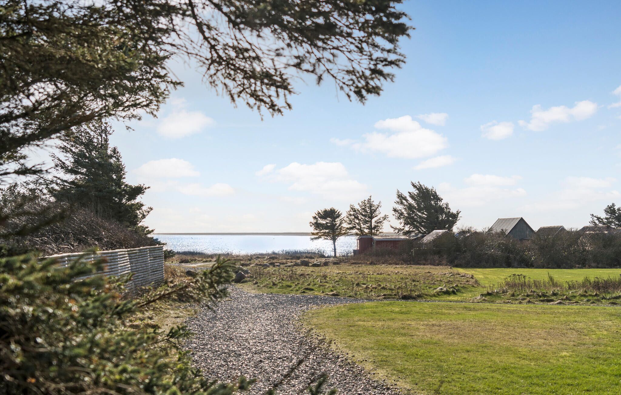 A coastal landscape with a gravel path leading to a wooden structure, surrounded by trees and a clear sky.