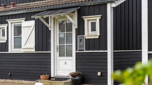 Een zwart-wit huis met een witte deur, twee ramen en een klein portaal met potplanten.