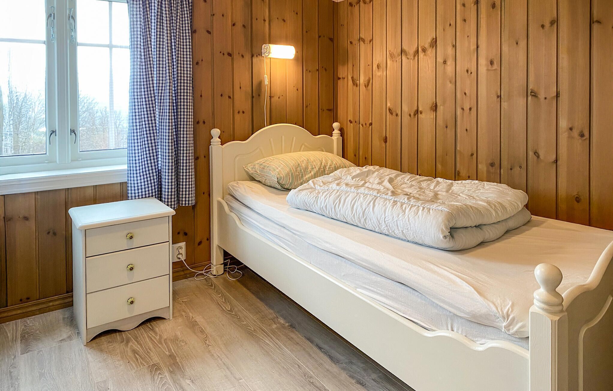 A bedroom with a wooden paneled wall, a white bed with a quilt, a bedside table with drawers, and a window with curtains.