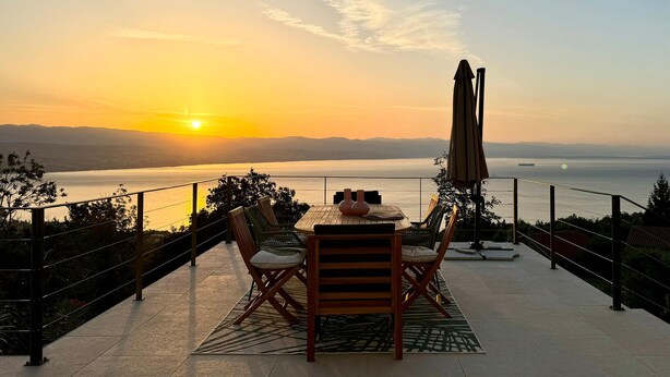 A terrace with a table and chairs facing a sunset over a body of water.