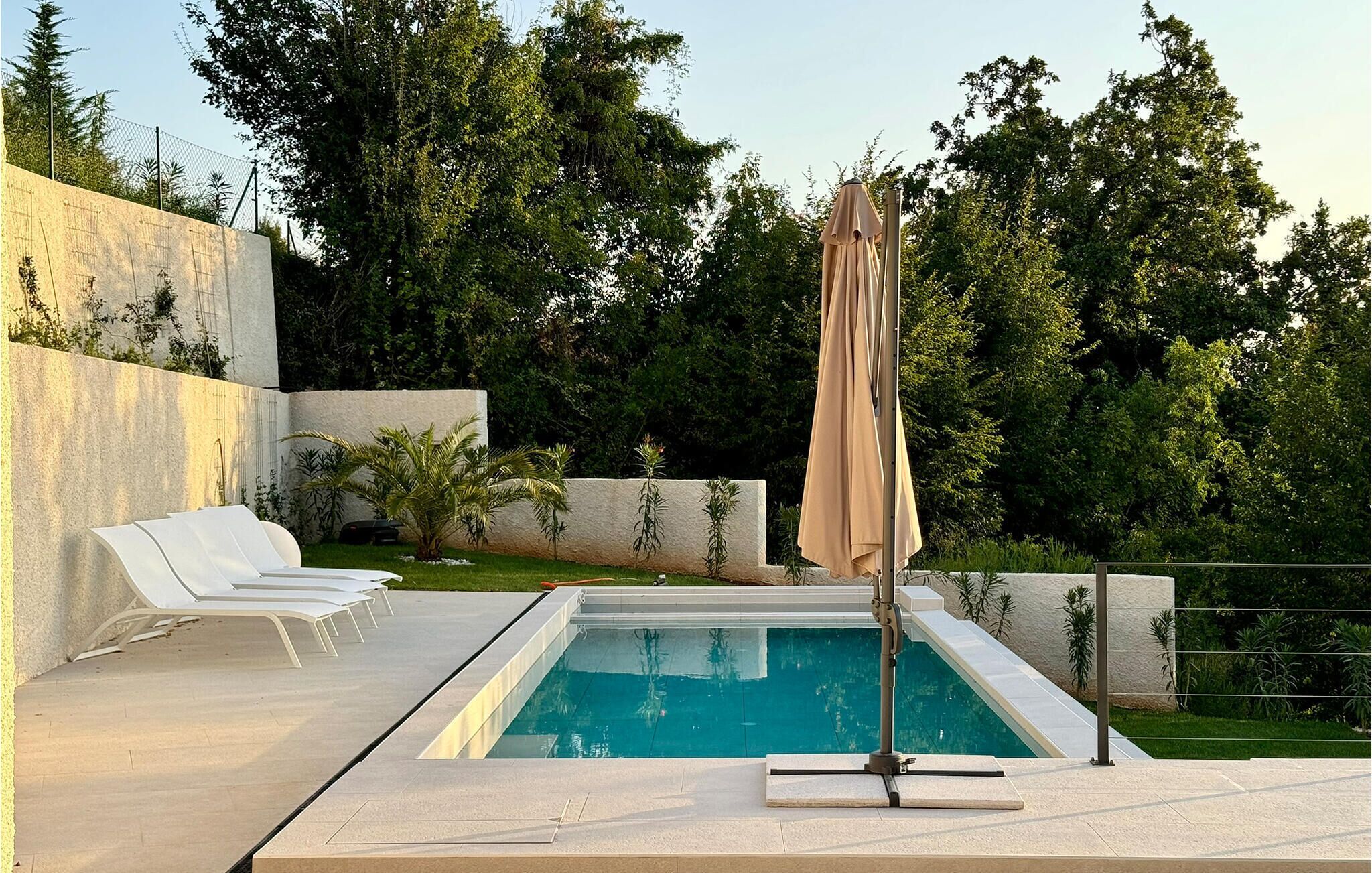 A modern outdoor pool area with a concrete deck, white lounge chairs, and a beige umbrella.