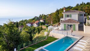 A modern villa with a swimming pool, green shutters, and a view of the sea.