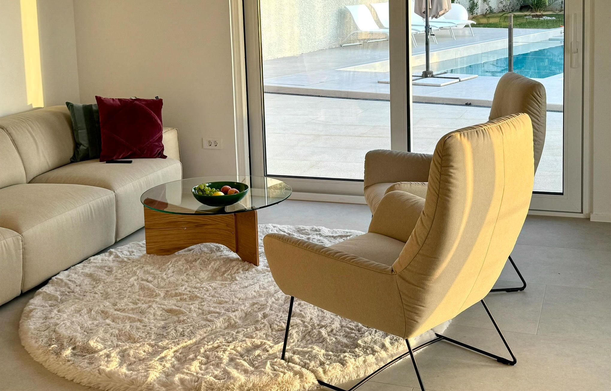 Un salon moderne avec un canapé beige, une table basse ronde en verre sur laquelle repose un saladier de fruits, un tapis blanc moelleux et un fauteuil beige.