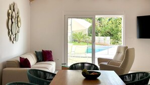 A living room with a sofa, chairs, a wooden table with a bowl of fruit, a TV, and a sliding glass door leading to a pool area.