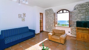 A living room with a blue sofa, a tan leather chair, a stone wall, a large window with a view, and a television on a wooden stand.