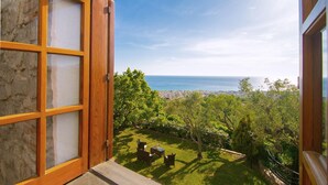 An open wooden door leading to a balcony with a view of the sea and a grassy area with outdoor furniture.