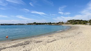 Une plage à l’eau d’un bleu limpide, avec un rivage sablonneux et, au loin, une ville où l’on aperçoit des bâtiments et de la verdure.