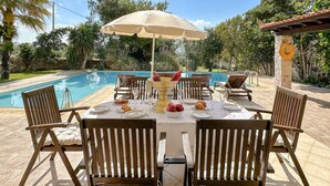 Un espace repas aménagé au bord de la piscine, avec une table dressée pour quatre personnes, entourée de chaises en bois, sous un parasol beige.