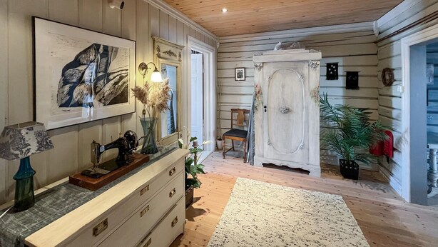 A room with a wooden ceiling, a white dresser with drawers, a sewing machine, a framed artwork, a chair, and a large white door. - Nice home in Sjusjøen with WiFi (Sjusjøen)