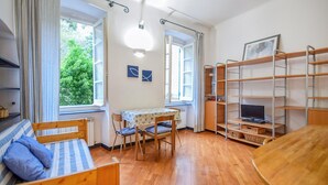 Ein Zimmer mit einem Bett, einem Tisch mit Stühlen, einem Fernseher auf einem Regal und großen Fenstern mit Vorhängen.