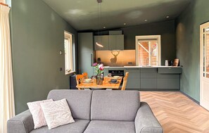 A modern living room with a grey sofa, a dining table with chairs, and a kitchen area with grey cabinets and a window. - Cozy home in Putten with WiFi (Putten)