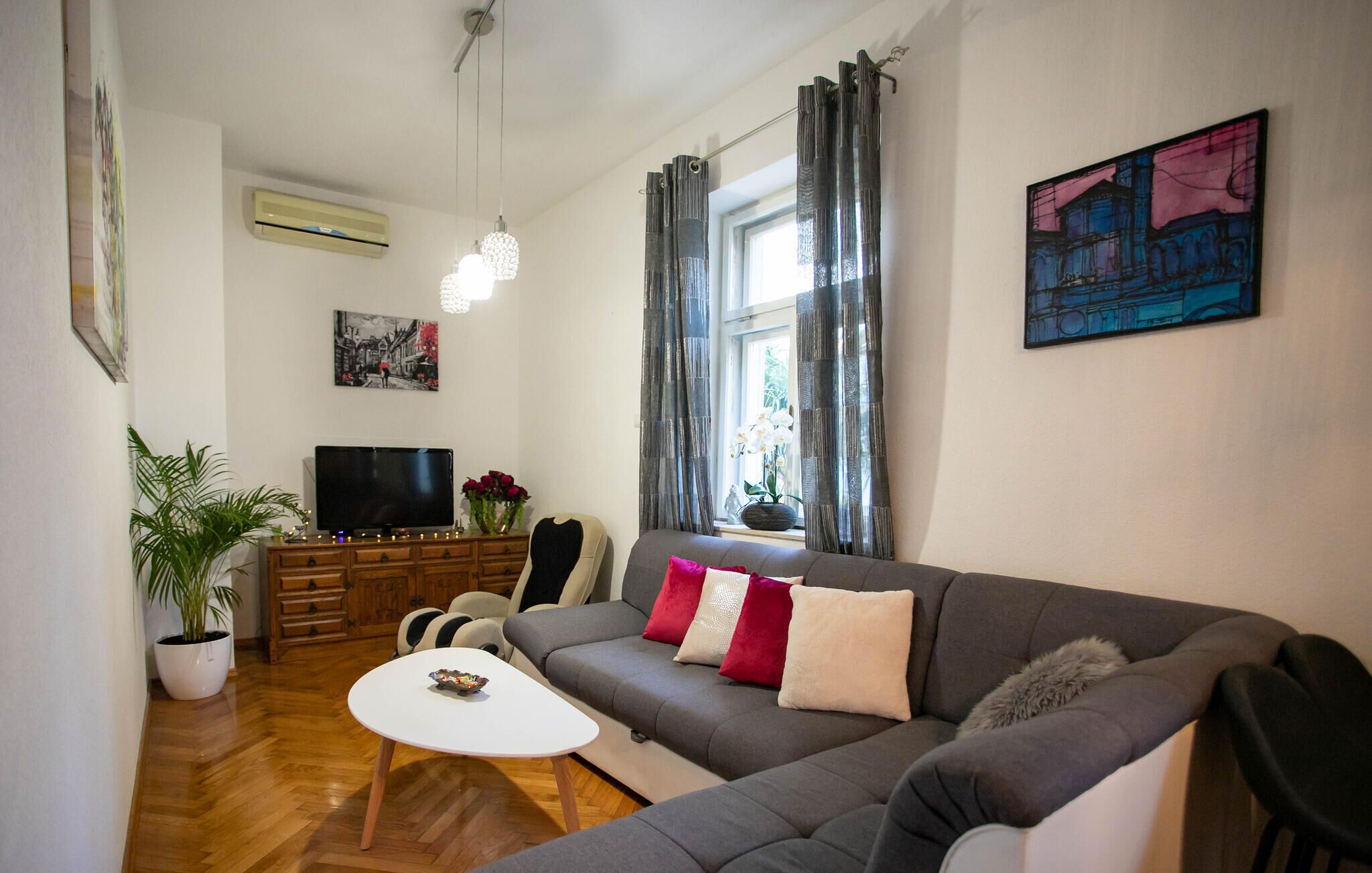 Sala de estar com sofá cinzento, mesa de centro branca, televisão, planta em vaso e um quadro na parede.