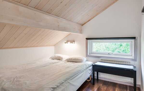 A bedroom with a sloped ceiling, a bed with pillows, a bench, and a window with a view of greenery.