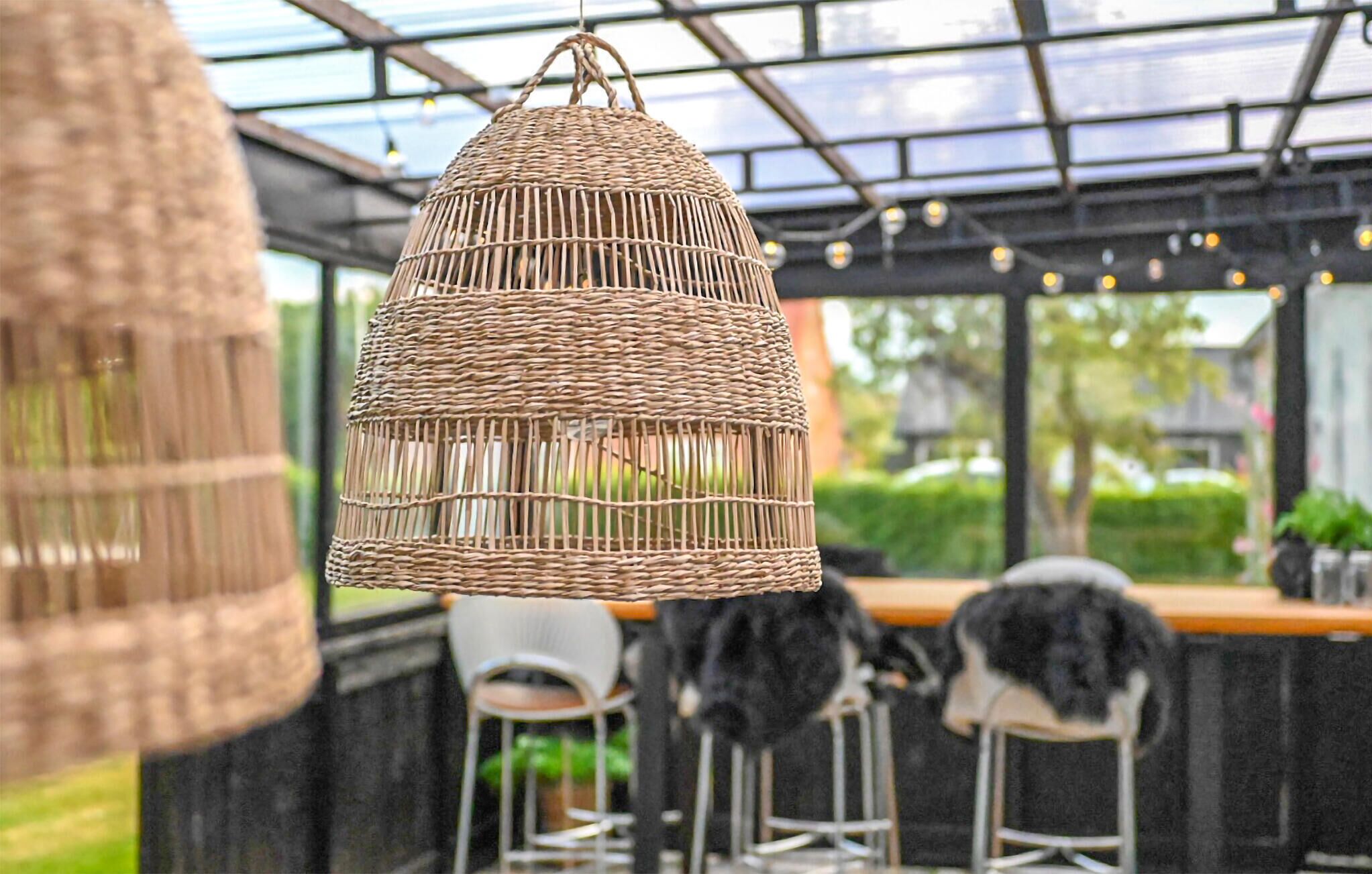 En hängande lampa av flätat material i ett rum som liknar ett växthus, med ett glastak, metallstolar och en bardisk.