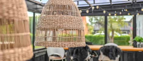 Un lampadario in vimini sospeso in una stanza in stile serra, con soffitto in vetro, sedie in metallo e un angolo bar.