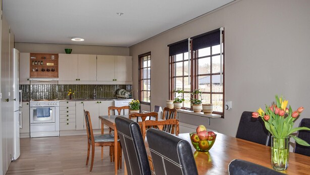 Una cocina moderna con armarios blancos, una mesa de comedor de madera y sillas. Hay ventanas con persianas, un lavaplatos y una estufa.