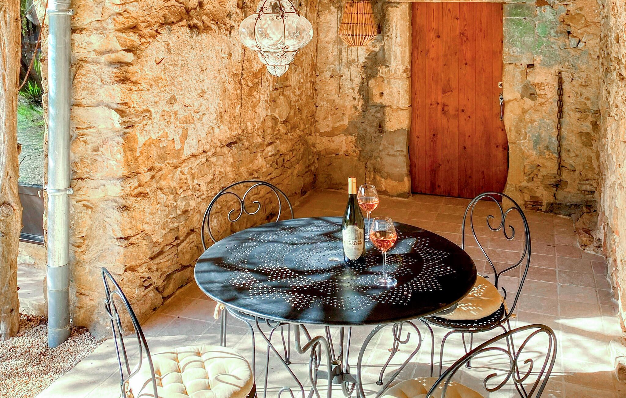 Un espace de restauration extérieur entouré de murs en pierre, avec une table ronde, des chaises en métal et des lanternes suspendues.