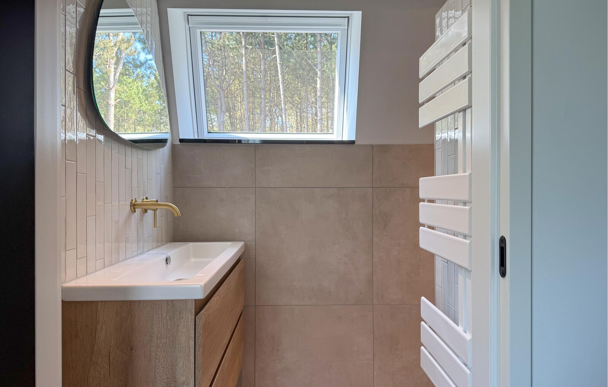 A bathroom with a white sink, wooden cabinetry, a gold faucet, and a window with a view of trees.