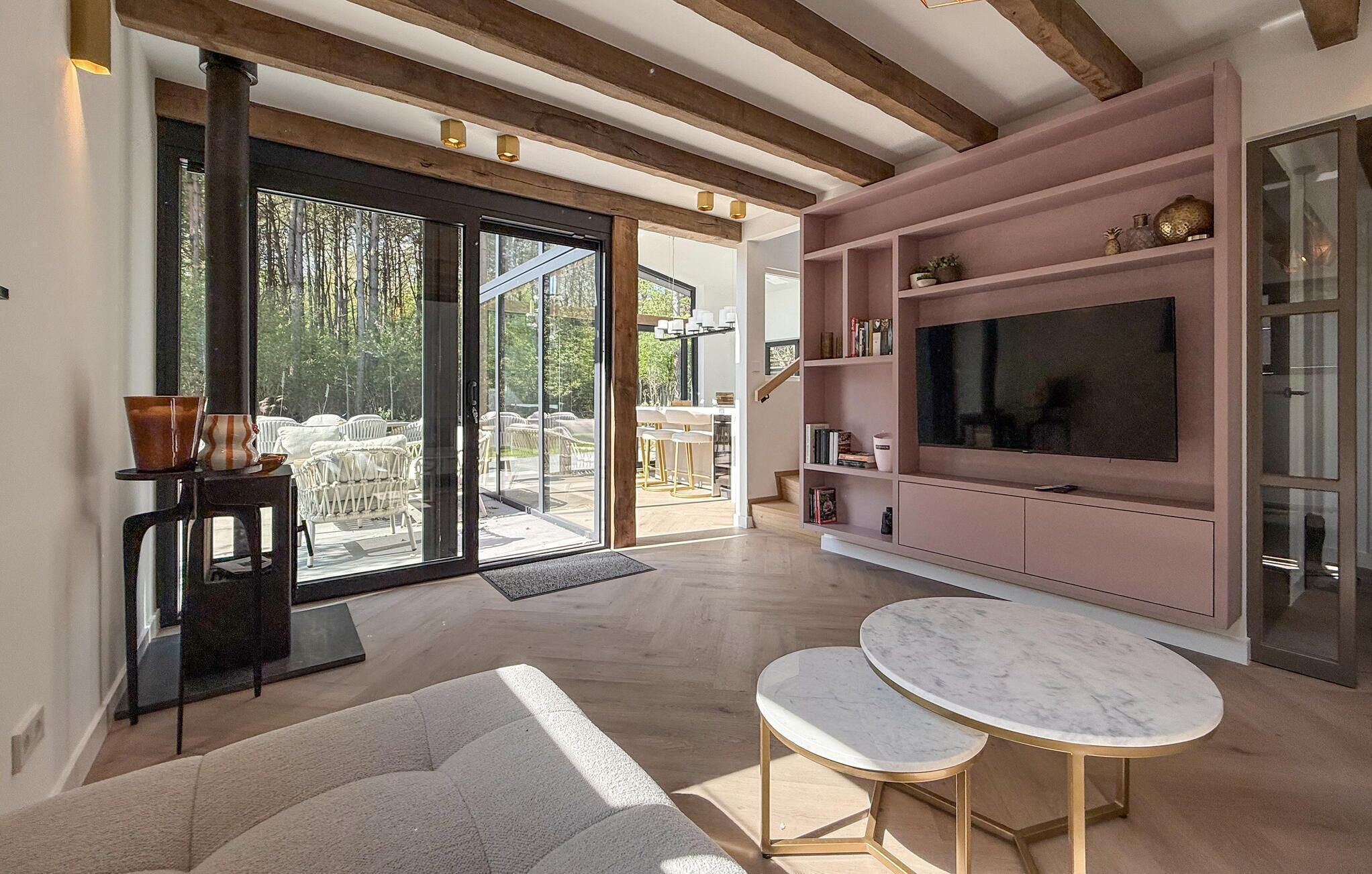 A modern living room with a large TV, wooden beams, a pink entertainment unit, and a glass door leading to an outdoor seating area.