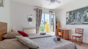 Une chambre d’hôtel avec deux lits, un bureau, une chaise, une grande fenêtre donnant sur des immeubles et un ventilateur de plafond.