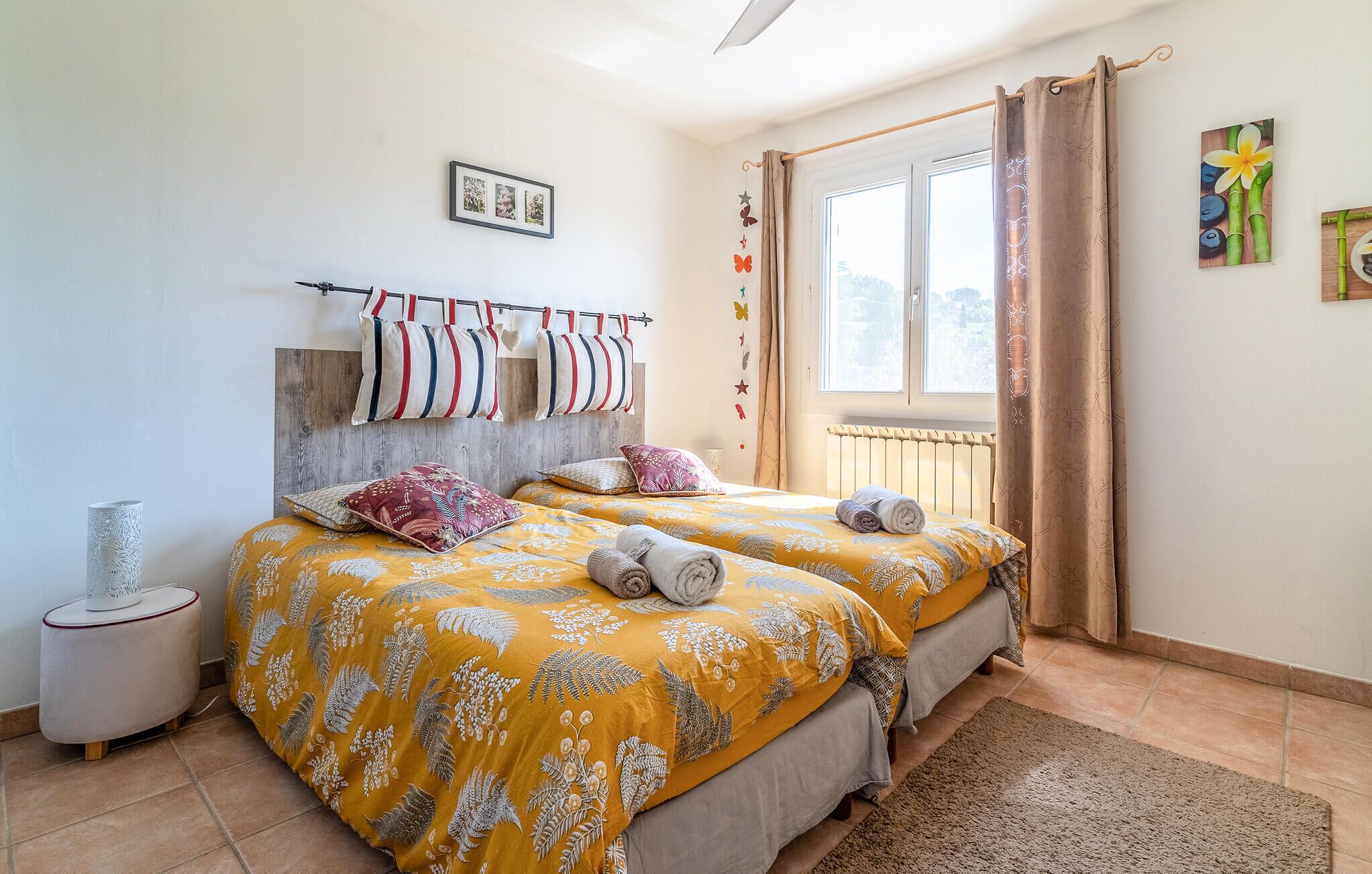Un dormitorio con dos camas, cabecero de madera, radiador, ventana con cortinas y una mesita auxiliar pequeña.