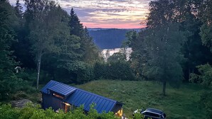 A cabin with a blue roof surrounded by dense forest, a car parked in front, and a view of a lake at sunset. - Cozy home in Tollered with WiFi (Tollered)