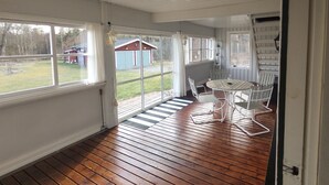 Eine überdachte Veranda mit Holzboden, einem weißen Tisch und Stühlen sowie Blick auf einen Garten mit einem Schuppen.