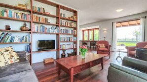 Ein geräumiges Wohnzimmer mit einer großen Bücherwand, einem Flachbildfernseher, einem Couchtisch und Blick auf eine grüne Landschaft durch große Fenster.